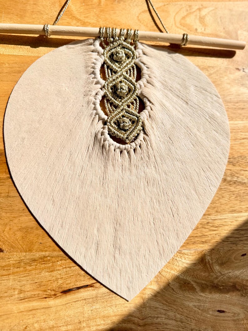Pode incluir: Um pendurador de parede bege em forma de folha com um design de macram&eacute; dourado. O macram&eacute; &eacute; preso a um pino de madeira. O pendurador de parede &eacute; feito de um material macio e texturizado, suspenso por um fino cord&atilde;o bege.