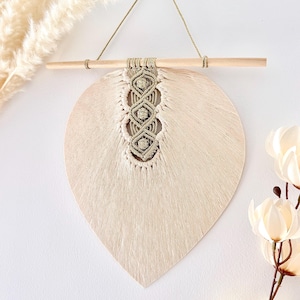 Pode incluir: Um pendurador de parede bege em forma de folha com um design de macram&eacute; no centro. A decora&ccedil;&atilde;o de parede &eacute; suspensa por um pino de madeira com um cord&atilde;o natural. A pe&ccedil;a &eacute; de cor neutra e complementaria qualquer decora&ccedil;&atilde;o.