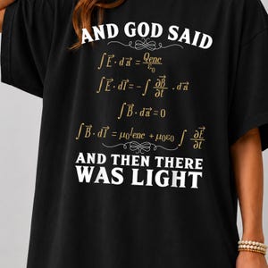 May include: A black t-shirt featuring white text that reads AND GOD SAID at the top and AND THEN THERE WAS LIGHT at the bottom. Between the text are Maxwell's equations printed in gold, including integral forms of Gauss's law and Faraday's law.