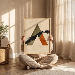 Puede incluir: Una impresión artística enmarcada con un diseño abstracto. La obra de arte presenta un gran triángulo verde, un triángulo naranja y una forma negra en forma de nube sobre un fondo beige. El marco es de madera clara. La impresión la sostiene una persona sentada en un suelo de madera.