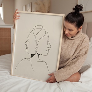 Könnte beinhalten: Ein rechteckiges gerahmtes Wandbild mit einer schwarzen durchgehenden Linienzeichnung von zwei Gesichtern im Profil, die Rücken an Rücken angeordnet sind. Das Kunstwerk befindet sich auf einem hellbeigen Hintergrund und der Rahmen ist aus hellem Holz.