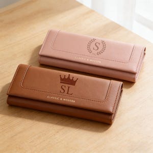 以下が含まれることがあります： ライトピンクとブラウンの2つの革製財布。ピンクの財布には月桂樹の冠の中に文字「S」があり、ブラウンの財布には王冠の下に「SL」が表示されています。どちらの財布にも「CLASSIC & MODERN」の文字があります。