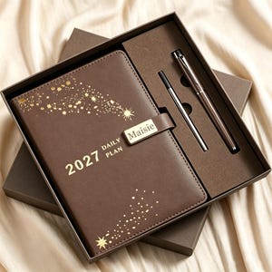 以下が含まれることがあります： 金色の星模様とMaisieと書かれたマグネット式留め具が付いた、茶色の2027年デイリープランナー。プランナーは同色の茶色のギフトボックスに入っており、茶色のボールペンと銀色のインク替え芯がセットになっています。