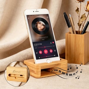 以下が含まれることがあります： 音楽インターフェースを表示するスマートフォンを置いた竹製の電話スタンド。スタンドには電話用のスロットと小さなスピーカーがあります。ペン立てや装飾品も表示されています。