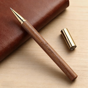 以下が含まれることがあります： 金色のペン先とアクセントが付いた木製のペンで、バレルには「Jeremy」という名前が刻まれています。 ペンの隣には、茶色の革のノートと金のペンキャップがあります。