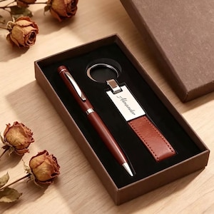 以下が含まれることがあります： 茶色のペンと、名前「Alexander」が銀色のプレートに刻まれたキーホルダーがセットになったギフトセット。ペンとキーホルダーは、黒い内装の茶色のギフトボックスに入っています。