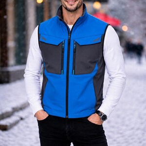 Può includere: Un gilet blu e grigio con cerniera a tutta lunghezza, due tasche sul petto e collo alto. Il gilet è indossato sopra una camicia bianca a maniche lunghe e abbinato a jeans neri. Un orologio è visibile sul polso sinistro.