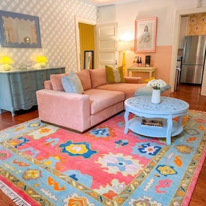 May include: A living room featuring a pink sectional sofa, a light blue round coffee table with a lower shelf, and a large area rug with a vibrant pink, light blue, yellow, and orange floral pattern. A blue dresser and a mirror are nearby.