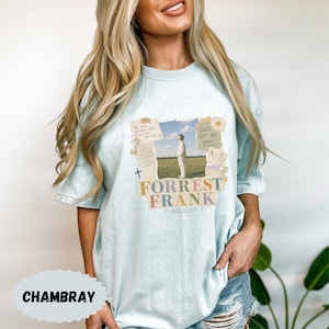 May include: A light blue t-shirt featuring a graphic print of a person standing in a field. The design includes torn paper-style borders, text reading "Be still and know that I am God," "GOD'S GOT MY BACK," "FORREST FRANK," and "child of God."