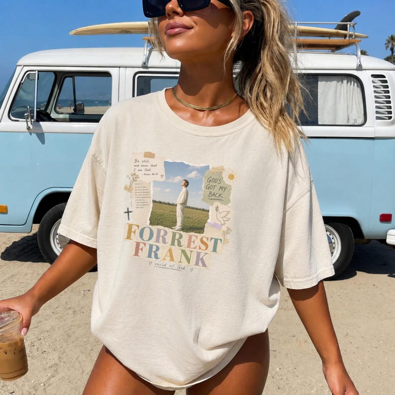 May include: A beige short-sleeve t-shirt featuring a graphic print with a photograph of a person standing in a field. The text reads Forrest Frank, God's Got My Back, and Be still, and know that I am God. The design includes a cross and a dove.