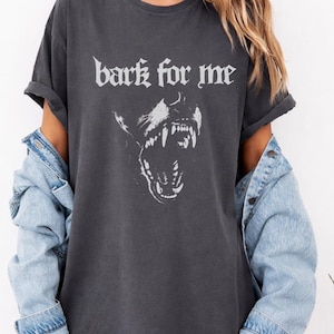 May include: Dark grey t-shirt featuring the text "bark for me" and a graphic of a snarling dog. The sleeves are rolled up. A denim jacket is worn over the shirt.