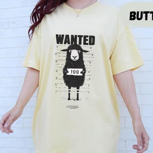 May include: Butter-colored t-shirt featuring a black sheep mugshot design with the word "WANTED" above it. The sheep has a number "100" on a sign around its neck. The shirt is a relaxed fit.