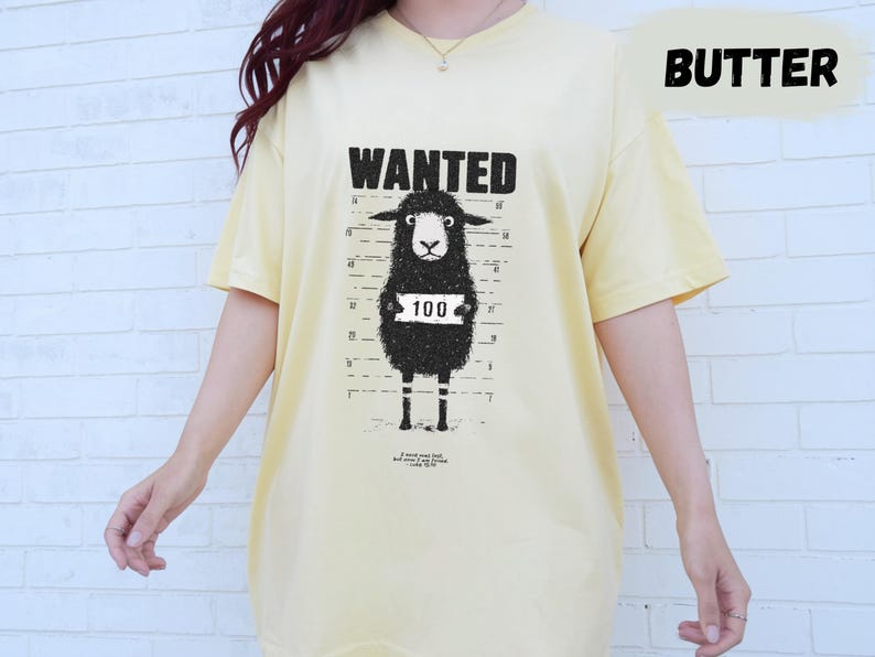 May include: Butter-colored t-shirt featuring a black sheep mugshot design with the word "WANTED" above it. The sheep has a number "100" on a sign around its neck. The shirt is a relaxed fit.