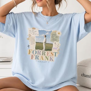 May include: A light blue t-shirt featuring a central graphic collage. The design includes a photograph of a person in a field, text reading "Be still and know that I am God," "God's got my back," "Forrest Frank," and "child of God," with a cross and dove.