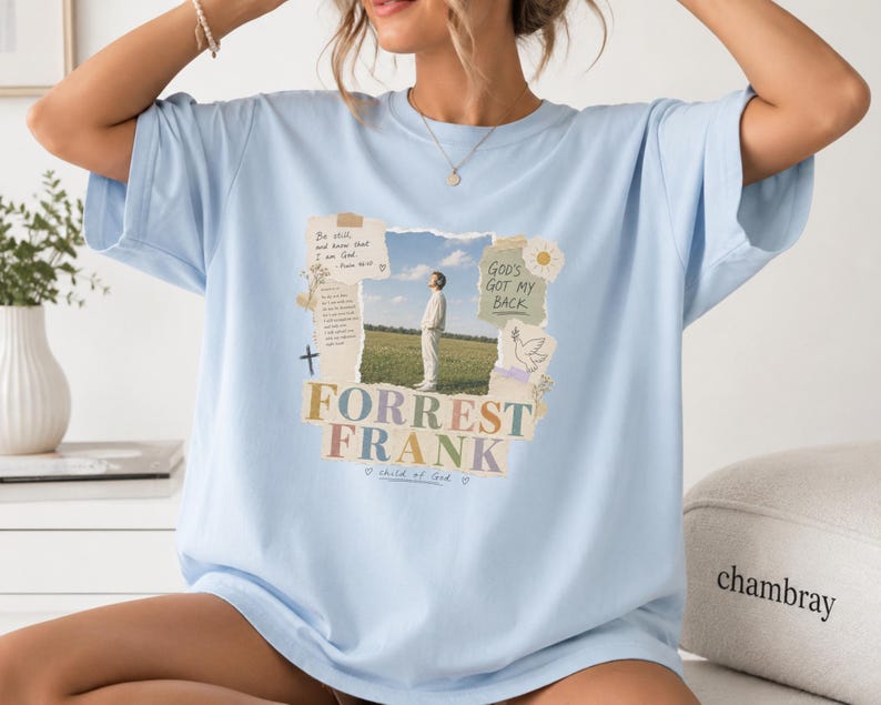May include: A light blue t-shirt featuring a central graphic collage. The design includes a photograph of a person in a field, text reading "Be still and know that I am God," "God's got my back," "Forrest Frank," and "child of God," with a cross and dove.