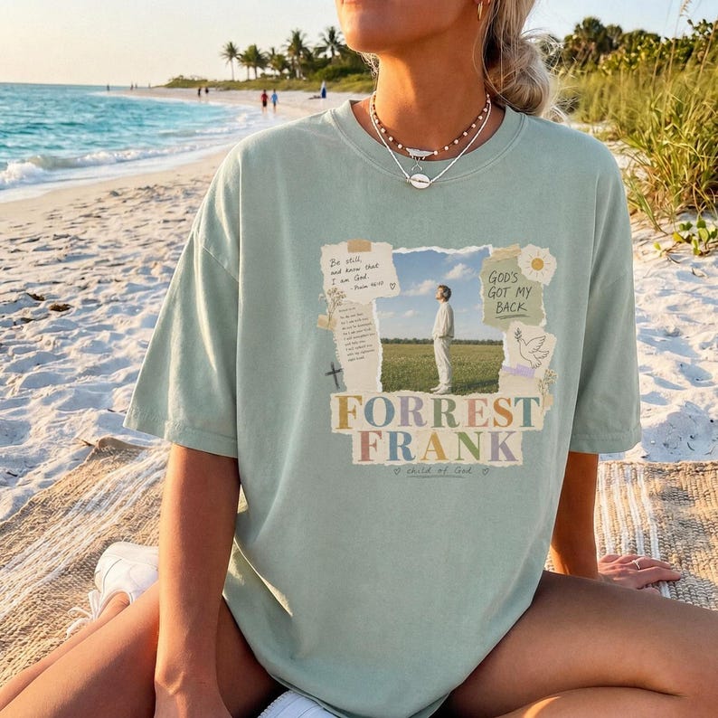 May include: A sage green short-sleeve t-shirt featuring a graphic print with a photograph of a person standing in a field, text that reads "Be still and know that I am God," "GOD'S GOT MY BACK," and "FORREST FRANK," plus a dove illustration.