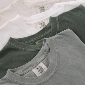 May include: Close-up of four Comfort Colors t-shirts in cream, white, olive green, and gray. Each shirt has a crew neck and a tag with the Comfort Colors logo. The shirts are folded and layered, showcasing their colors and textures.