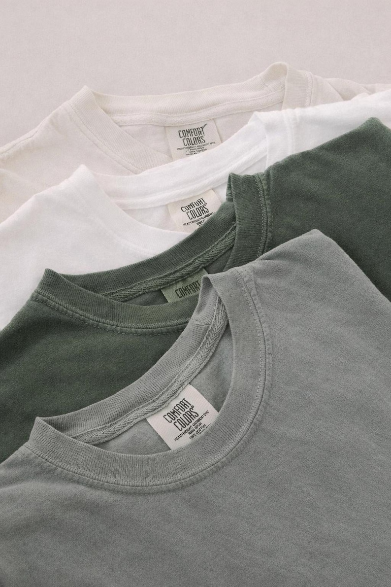May include: Close-up of four Comfort Colors t-shirts in cream, white, olive green, and gray. Each shirt has a crew neck and a tag with the Comfort Colors logo. The shirts are folded and layered, showcasing their colors and textures.