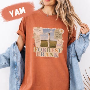 May include: A rust-coloured short-sleeve t-shirt featuring a collage-style graphic print. The design includes a photograph of a person in a field, text reading "Be still, and know that I am God," "GOD'S GOT MY BACK," and "FORREST FRANK," plus a cross and dove.
