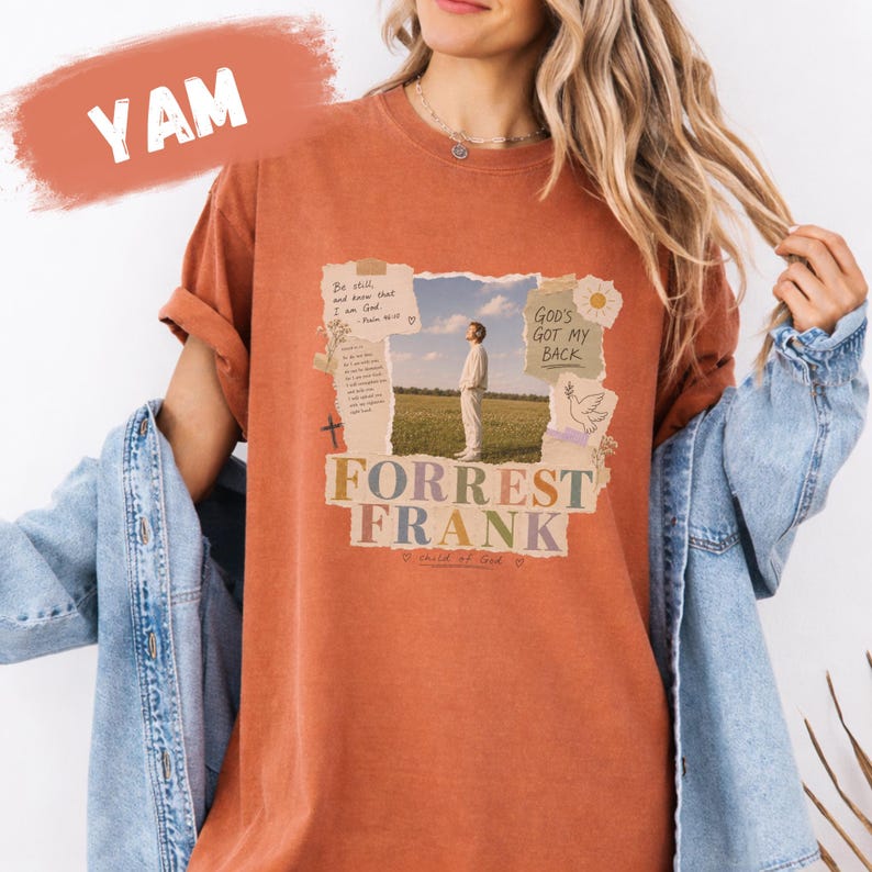 May include: A rust-coloured short-sleeve t-shirt featuring a collage-style graphic print. The design includes a photograph of a person in a field, text reading "Be still, and know that I am God," "GOD'S GOT MY BACK," and "FORREST FRANK," plus a cross and dove.