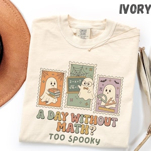May include: An ivory t-shirt featuring a graphic of three postage stamp-style illustrations of ghosts with maths-themed elements like books, a chalkboard, and geometry tools. The text reads A DAY WITHOUT MATH? TOO SPOOKY in a retro font.