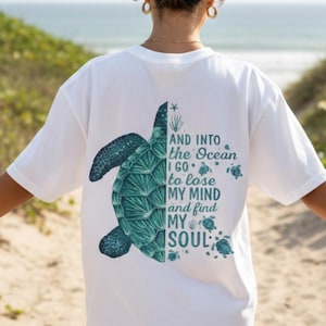 Puede incluir: Camiseta blanca con un diseño de tortuga marina turquesa y el texto "AND INTO the Ocean I go to lose MY MIND and find MY SOUL." El diseño incluye tortugas más pequeñas y elementos acuáticos.