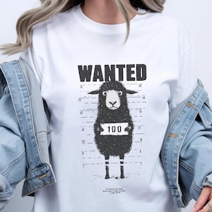 May include: White t-shirt with a black graphic of a sheep in a police lineup, holding a sign with the number 100. The word "WANTED" is above the sheep. The shirt also has a Bible verse at the bottom.