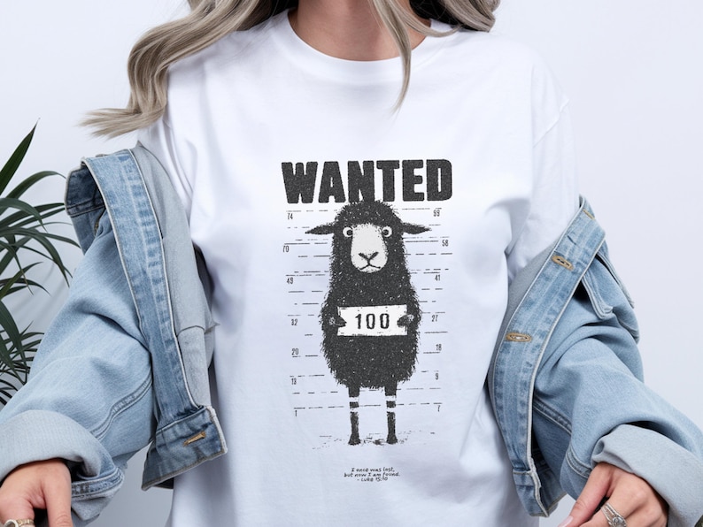 May include: White t-shirt with a black graphic of a sheep in a police lineup, holding a sign with the number 100. The word "WANTED" is above the sheep. The shirt also has a Bible verse at the bottom.