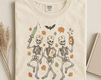 以下が含まれることがあります： アイボリー色のTシャツで、看護師の格好をした3体の骸骨が描かれています。それぞれの骸骨はナースキャップを被っており、周囲にはオレンジ色の秋の葉、コウモリ、注射器、クリップボード、薬瓶が配置されています。