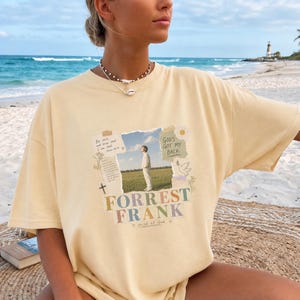 May include: A pale yellow short-sleeve t-shirt featuring a central graphic print. The design includes a photograph of a person in a field, surrounded by text that reads "Be still, and know that I am God," "GOD'S GOT MY BACK," and "FORREST FRANK."