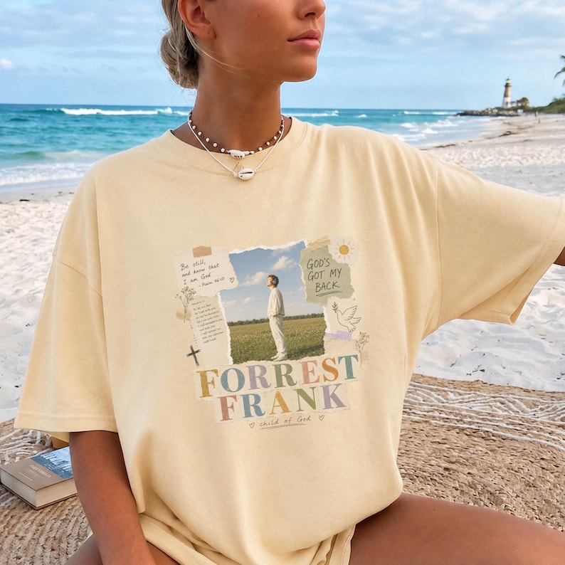 May include: A pale yellow short-sleeve t-shirt featuring a central graphic print. The design includes a photograph of a person in a field, surrounded by text that reads "Be still, and know that I am God," "GOD'S GOT MY BACK," and "FORREST FRANK."