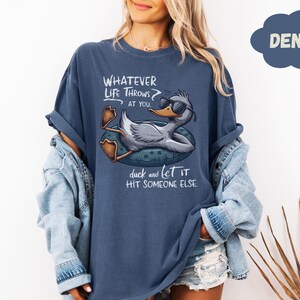 Op de afbeelding: Denim blauw T-shirt met een afbeelding van een eend met een zonnebril die op een zwemband drijft. De tekst luidt: "Wat het leven je ook toewerpt, duik en laat het iemand anders raken."