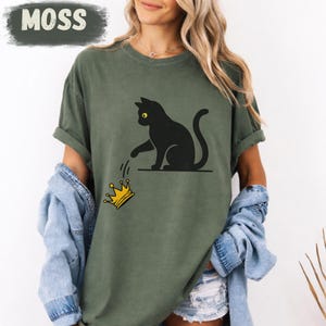 Pode incluir: Camiseta verde-oliva com um gato preto e uma coroa dourada. O gato tem olhos amarelos. A palavra "MOSS" está impressa em um retângulo verde escuro no canto superior esquerdo da camiseta.