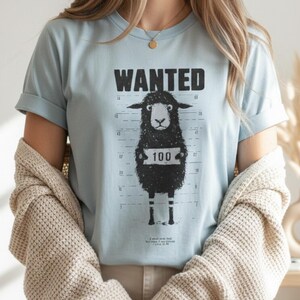 May include: Light blue t-shirt with the word "WANTED" above a black sheep holding a sign that says "100". The shirt has a mugshot-style design with a height chart in the background.