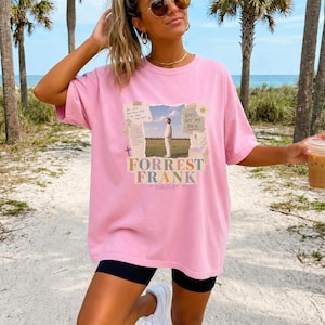 May include: A light pink short-sleeve t-shirt featuring a central graphic print. The design includes a photograph of a person in a field, surrounded by torn paper elements, a dove, and text that reads Forrest Frank, God's Got My Back, and Child of God.