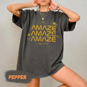 Könnte beinhalten: Dunkelgraues T-Shirt mit dem Wort "AMAZE" dreimal in Gold, mit einem Planeten- und Sternen-Motiv. Der Text "Ryland and Rocky" steht darunter. Das Shirt hat die Farbe Pfeffer.
