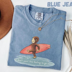 May include: Blue jean colored t-shirt with a graphic of a monkey surfing on a red surfboard. The text "I don't want this feeling to go away" is printed at the bottom. A gold necklace is draped across the shirt.