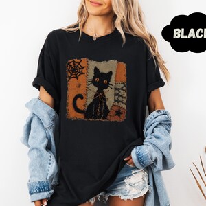 May include: A black t-shirt featuring a square graphic print of a black cat with orange eyes, surrounded by orange and brown patchwork-style patterns, a spider web, and a star. The word BLACK is printed in a black cloud shape in the corner.