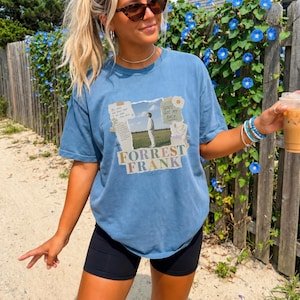 May include: A blue short-sleeve t-shirt featuring a central graphic print of a person standing in a field. The design includes torn paper elements, text reading "FORREST FRANK" and "GOD'S GOT MY BACK," a small dove illustration, and additional printed text.