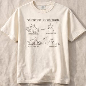 Puede incluir: Camiseta color crema con el texto "SCIENTIFIC MEOWTHOD" sobre un diagrama del proceso científico de un gato. El diagrama muestra a un gato observando, formulando una hipótesis, experimentando y analizando.