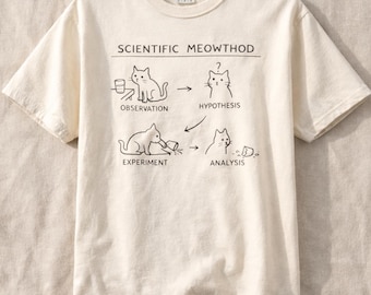 May include: Cream-colored t-shirt with the text "SCIENTIFIC MEOWTHOD" above a diagram of a cat's scientific process. The diagram shows a cat observing, forming a hypothesis, experimenting, and analyzing.