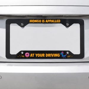 Puede incluir: Un marco de matrícula rectangular negro con el texto amarillo "MONGO IS APPALLED" en el borde superior y "AT YOUR DRIVING" en el borde inferior, acompañado de un gráfico de una rosquilla glaseada rosa y un pequeño dinosaurio azul.
