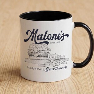 Puede incluir: Taza de cerámica blanca con interior y asa negros. La taza presenta un gráfico de una hamburguesa y patatas fritas, con el texto "Malone's since 2015" y "Proudly Serving Briar University" en azul marino.