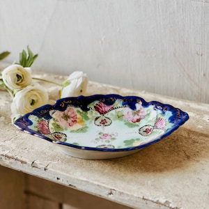 May include: An antique, oval ceramic dish with a scalloped edge. The dish is decorated with a floral pattern in pink, green, and red, set against a light blue background. The rim is a deep blue with gold detailing, adding a touch of elegance.