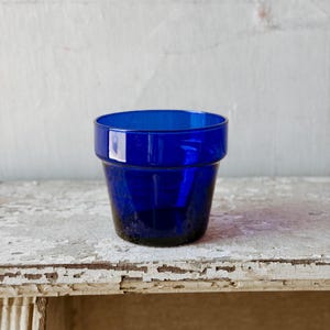 May include: A small, cylindrical cobalt blue glass vessel with a smooth, translucent finish. The object features a distinct, slightly flared rim at the top and a solid, flat base, showcasing a deep, vibrant blue hue throughout the glass structure.