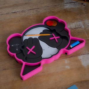 Può includere: Un originale organizer da scrivania rosa e nero a forma di personaggio dei cartoni animati. Presenta un volto bianco, mani nere con segni 'X' rosa e un vassoio contenente una matita, una spilla da balia e un oggetto blu. L'organizer è su una superficie di legno.