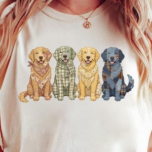 Pode incluir: Camiseta creme com um gráfico de quatro cães golden retriever. Cada cão usa um acessório estampado diferente: uma bandana, um lenço e dois lenços xadrez diferentes. Os cães estão sorrindo e o desenho está centralizado na camiseta.