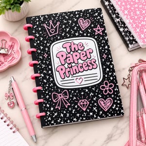 May include: A black disc-bound notebook with a white speckled pattern and pink accents. The cover features the text The Paper Princess in pink bubble letters, surrounded by illustrations of a crown, hearts, a star, a bow, a flower, and a diamond.