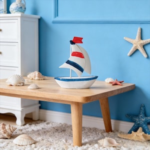 May include: A nautical-themed decorative sailboat with a white hull and blue trim, red and blue accents on the sail, and a red flag. The sailboat sits on a wooden table with seashells and starfish, against a blue wall.
