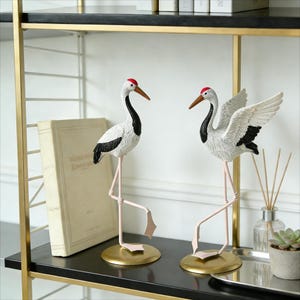 Könnte beinhalten: Zwei dekorative Kranichfiguren. Ein Kranich hat seine Flügel ausgebreitet. Die Vögel sind weiß mit schwarzen Akzenten und roten Kronen. Sie stehen auf goldfarbenen Sockeln mit rosa Beinen. Die Figuren stehen auf einem schwarzen Regal.